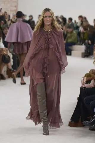 A model at the Chloé autumn/winter 2024 runway show wearing a sheer ruffled maxi dress with thigh-high boots and a gold necklace.