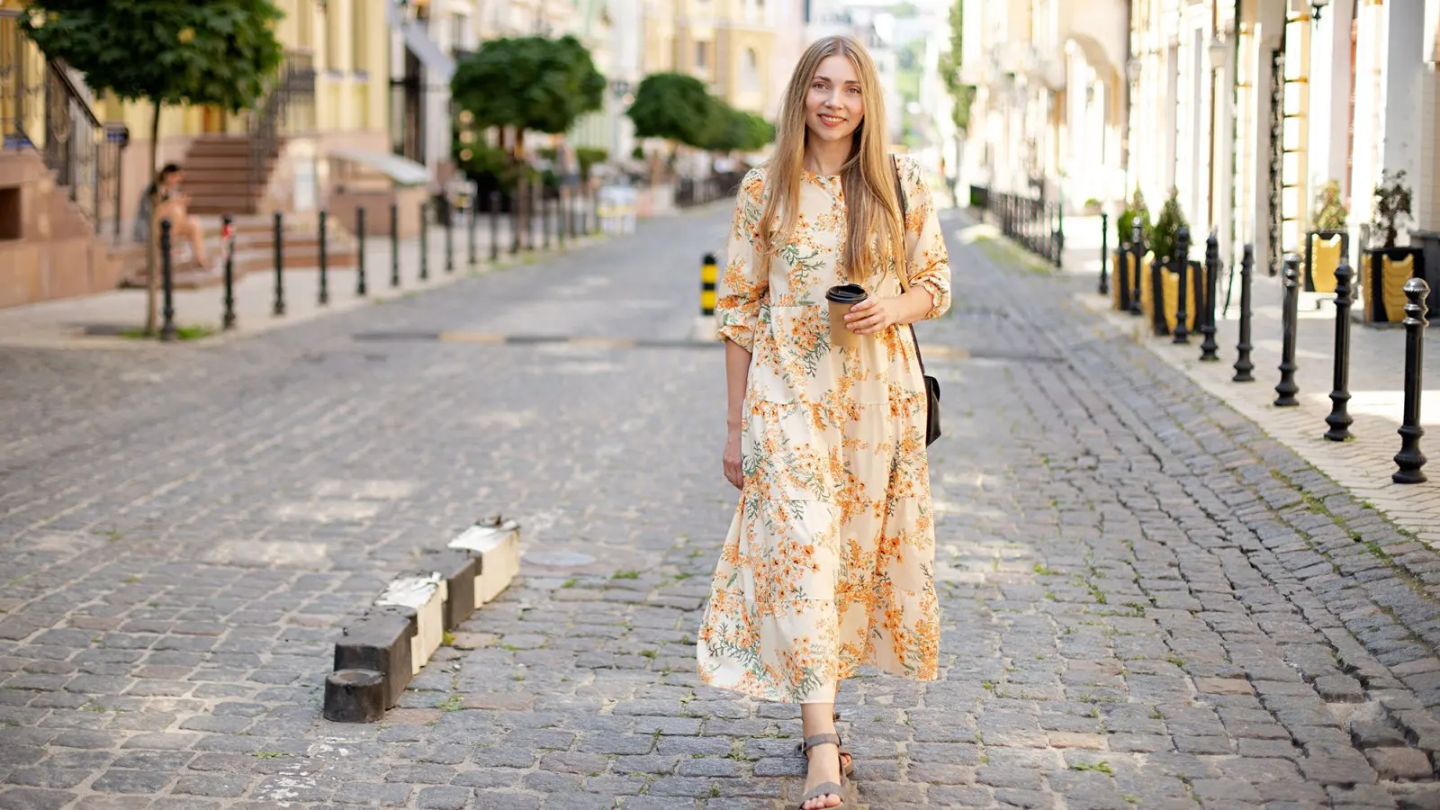 smiling woman blonde in dress with coffee takeaway walks on the cobblestones against the backdrop of the street and green trees in summer