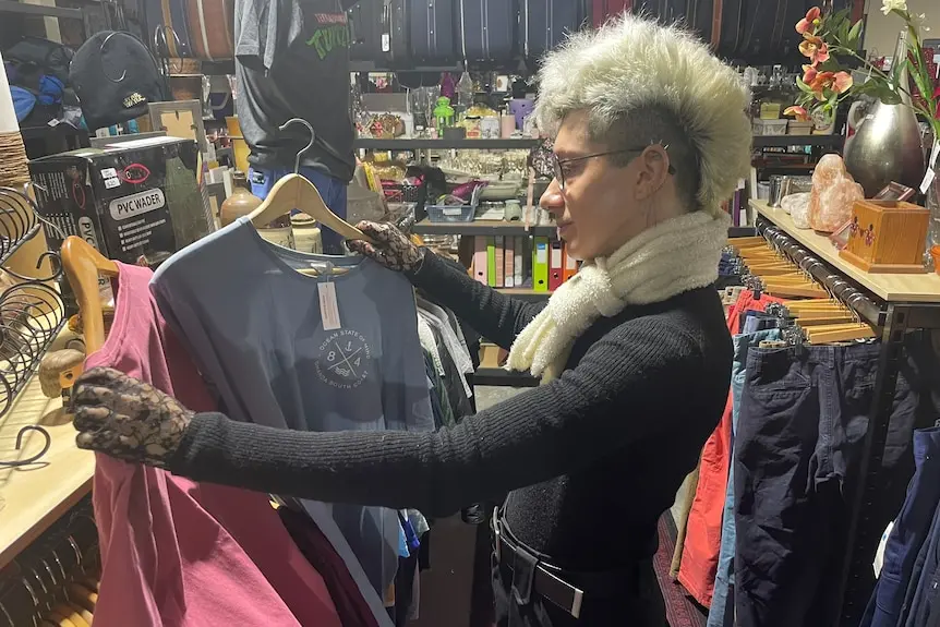 A person with a short blond mohawk hairstyle, black top and scarf compares two t-shirts in an op shop