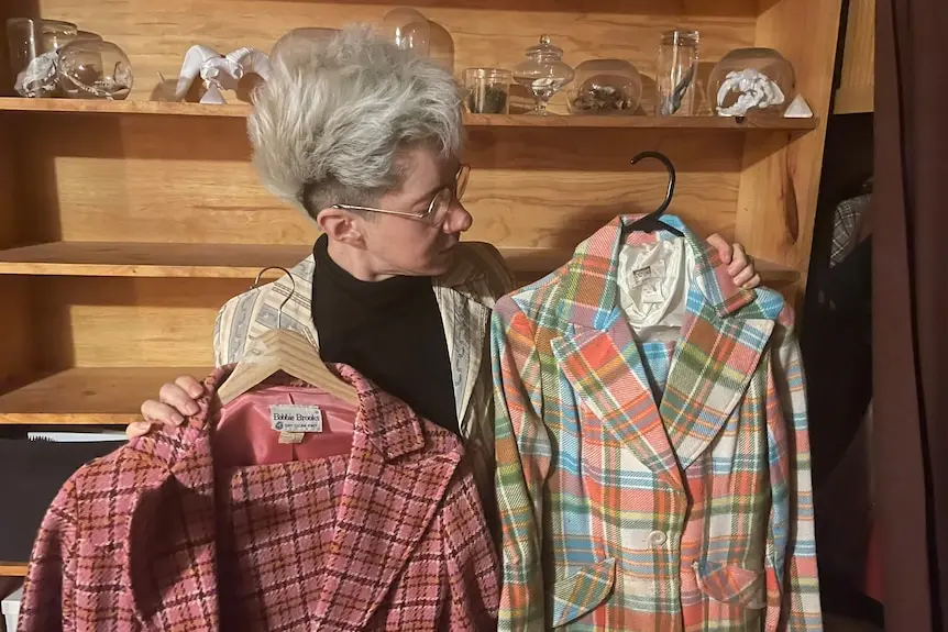 A person with a short blond mohawk hairstyle looks at two colourful patterned suits in a shop