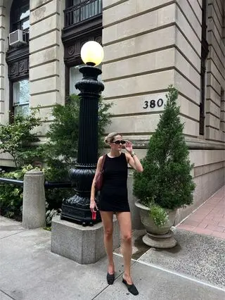 Eliza Huber wearing a black Norma Kamali Pickleball Dress with black mules, sunglasses, and a burgundy handbag.