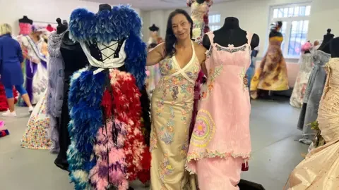 Tanys Pullin next to two dresses - one is a blue, red and pink dress covered in feathers. The other is a pink dress with some flowers and a yellow circle decorating it.