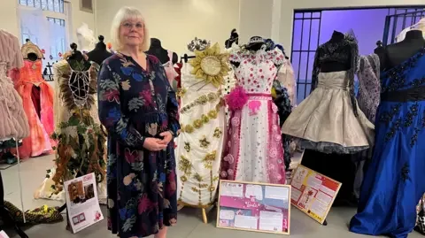 Marjorie Wells standing next to her dresses - one is white with gold flowers wrapped around it and a sunflower at the top. The other is white with pink flowers decorating it.