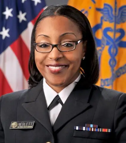 Doctor Robyn Neblett Fanfair is seen from the shoulders up. She is posing in front of a pair of flags and smiling. She has short dark hair and glasses. She is also wearing what looks like a black military jacket with pins on it.