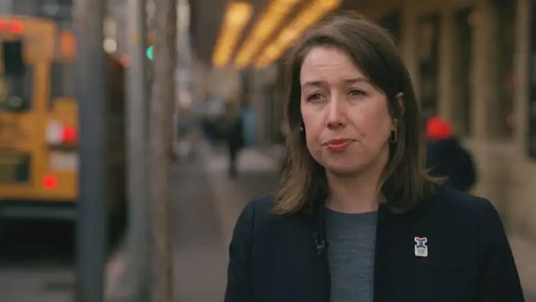 A woman stands on a sidewalk during an interview. She has brown shoulder-length hair. She wears a grey blouse and black blazer.