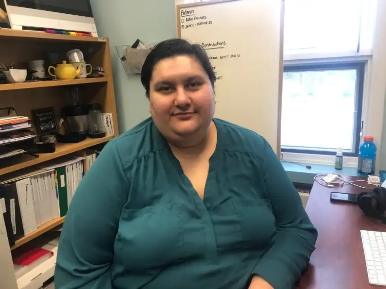 A person in a teal long-sleeve shirt sits at a desk and looks at the camera