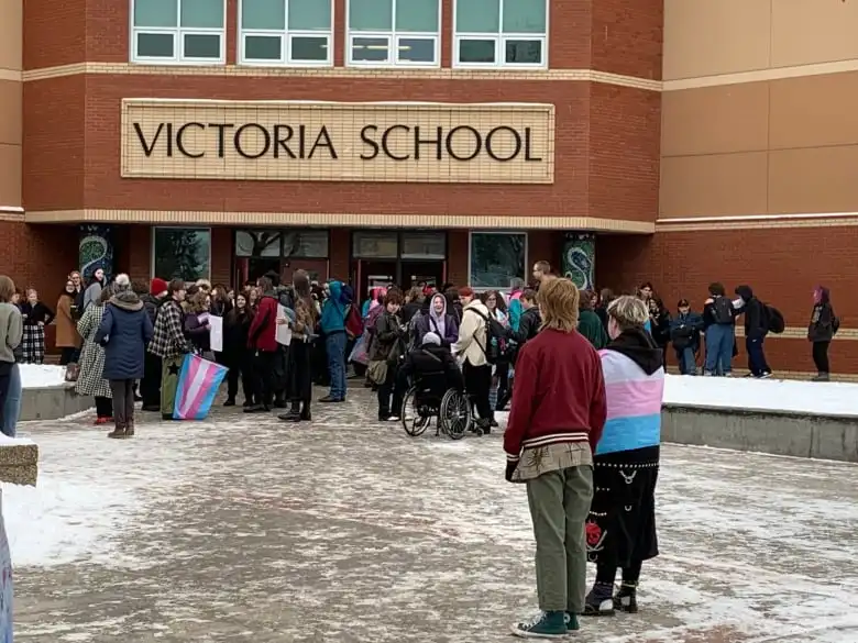 Students stand outside the doors of a brick building. The sign above the building's entry doors reads Victoria School.