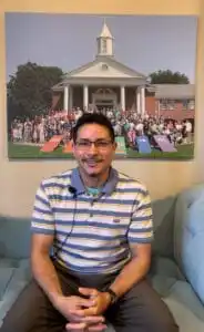 Saer Olavarria has brown, olive skin, with short dark hair. He wears rectangular shaped glasses, and has a mustache with a soul patch. He wears a blue and white striped polo shirt, with grey pants. He sits on a blue couch in Glendale United Methodist Church with a frame of members of the church behind him.