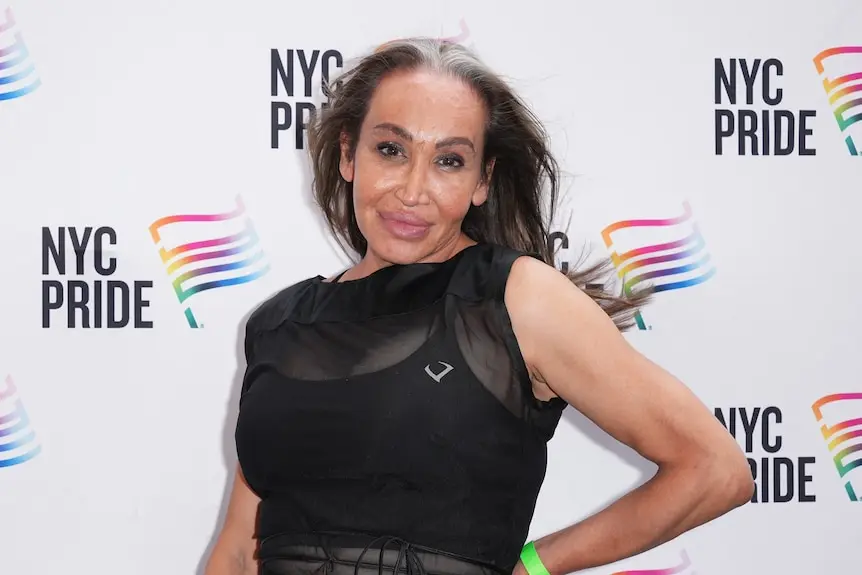 A close up of a woman with long hair standing and smiling in front of a wall with 'NYC Pride' logos all over it