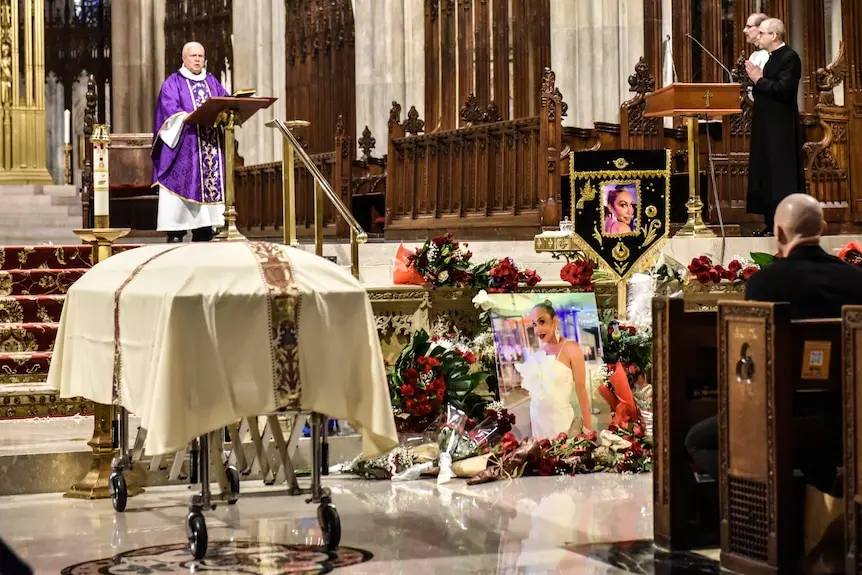 A wide shot of a priest speaking in a large church. People sitting in pews and standing nearby. A casket in the aisle.