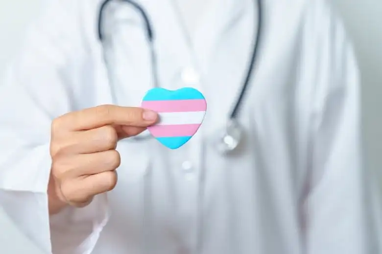 Stock image of a person wearing a lab coat and stethoscope, pictured from the shoulders down, holding out a small plastic heart with light blue, pink and white stripes — the colours of the transgender pride flag.