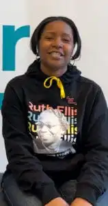 Sheree Lampinsky (she/her), a black woman with brown eyes, and dark brown hair tied back in a ponytail, sits with her hands on her knees wearing a Ruth Ellis sweatshirt that reads 