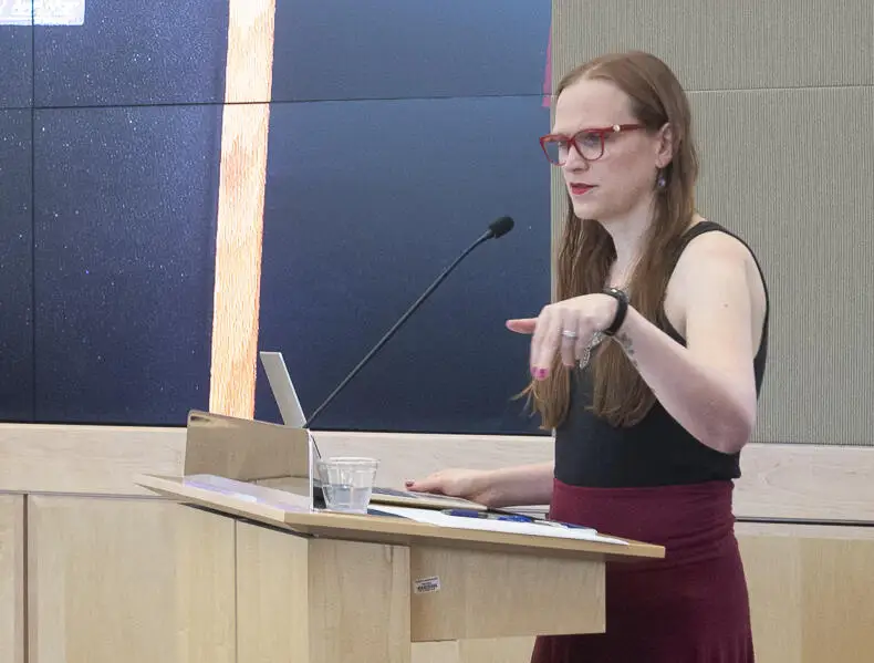 A photo of a woman standing a a podium and speaking.