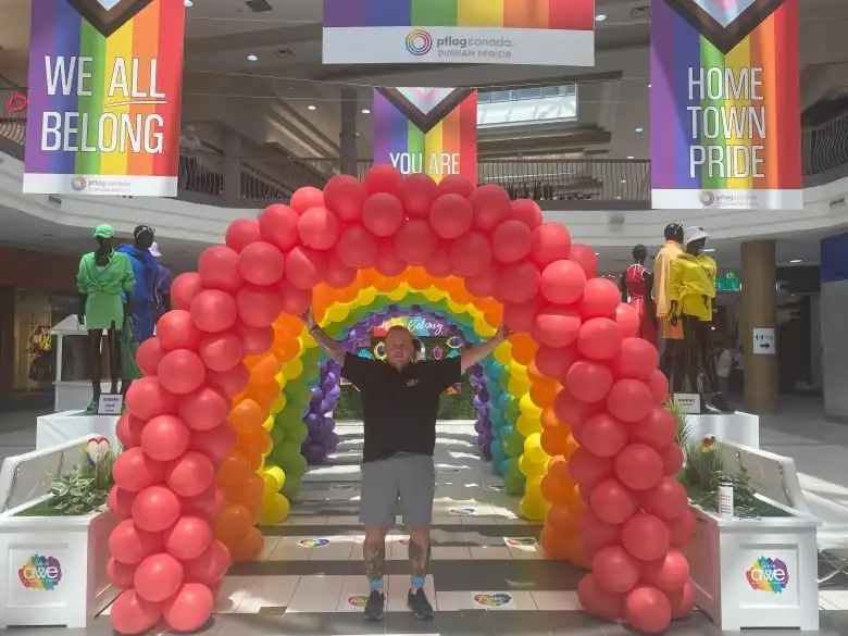 Farr stands beneath a rainbow made of balloons, part of a promotional display in a shopping mall.