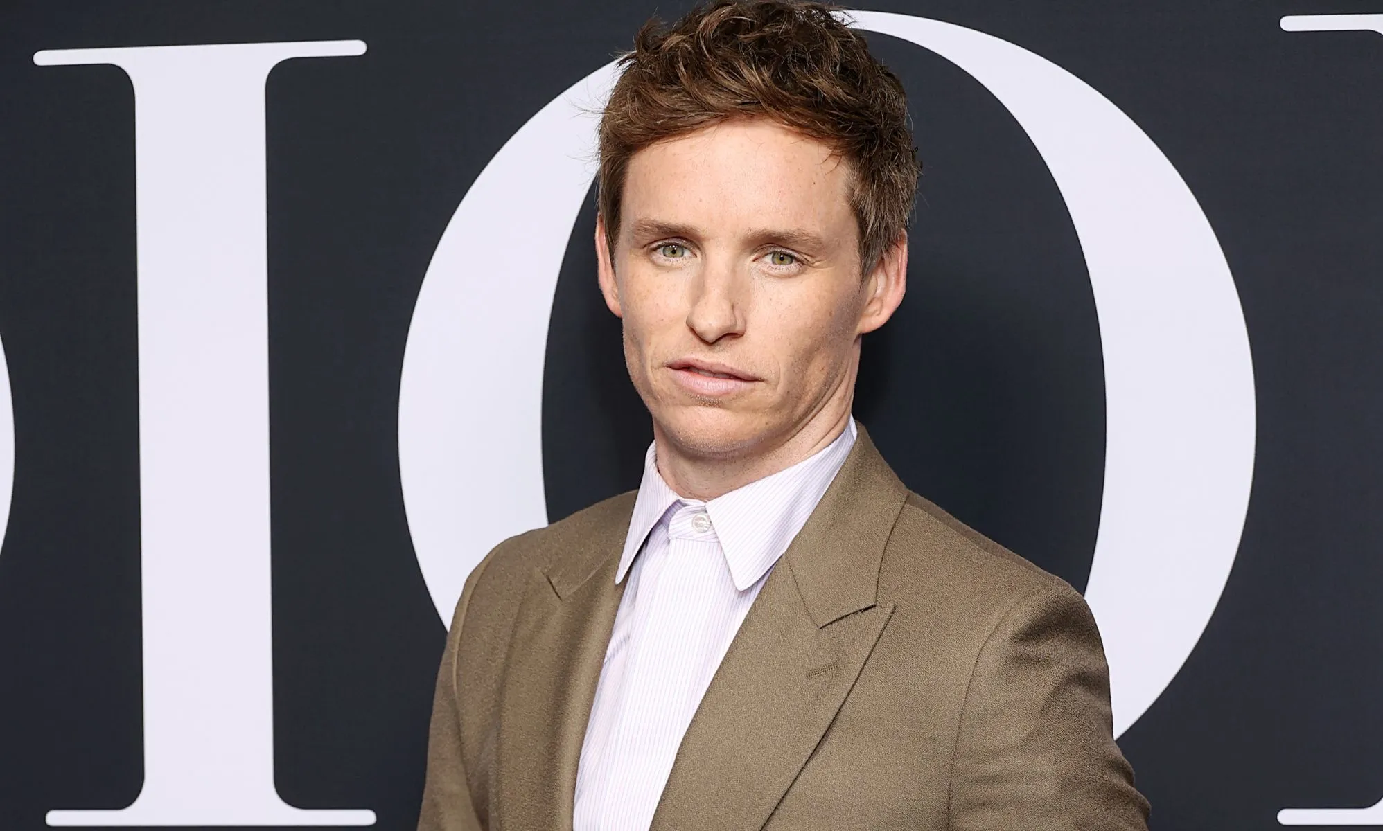 Eddie Redmaybe wears a white button-up shirt with a light brown jacket as he poses for the camera at an event