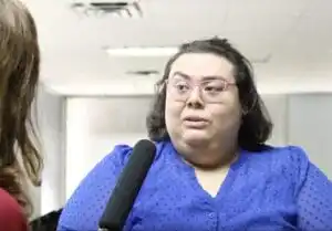 Gia Pacheco, transgender, immigration activist, with brown hair, fair complexion and glasses, wearing a blue dress, and pink, wing framed glasses, speaks into a black microphone