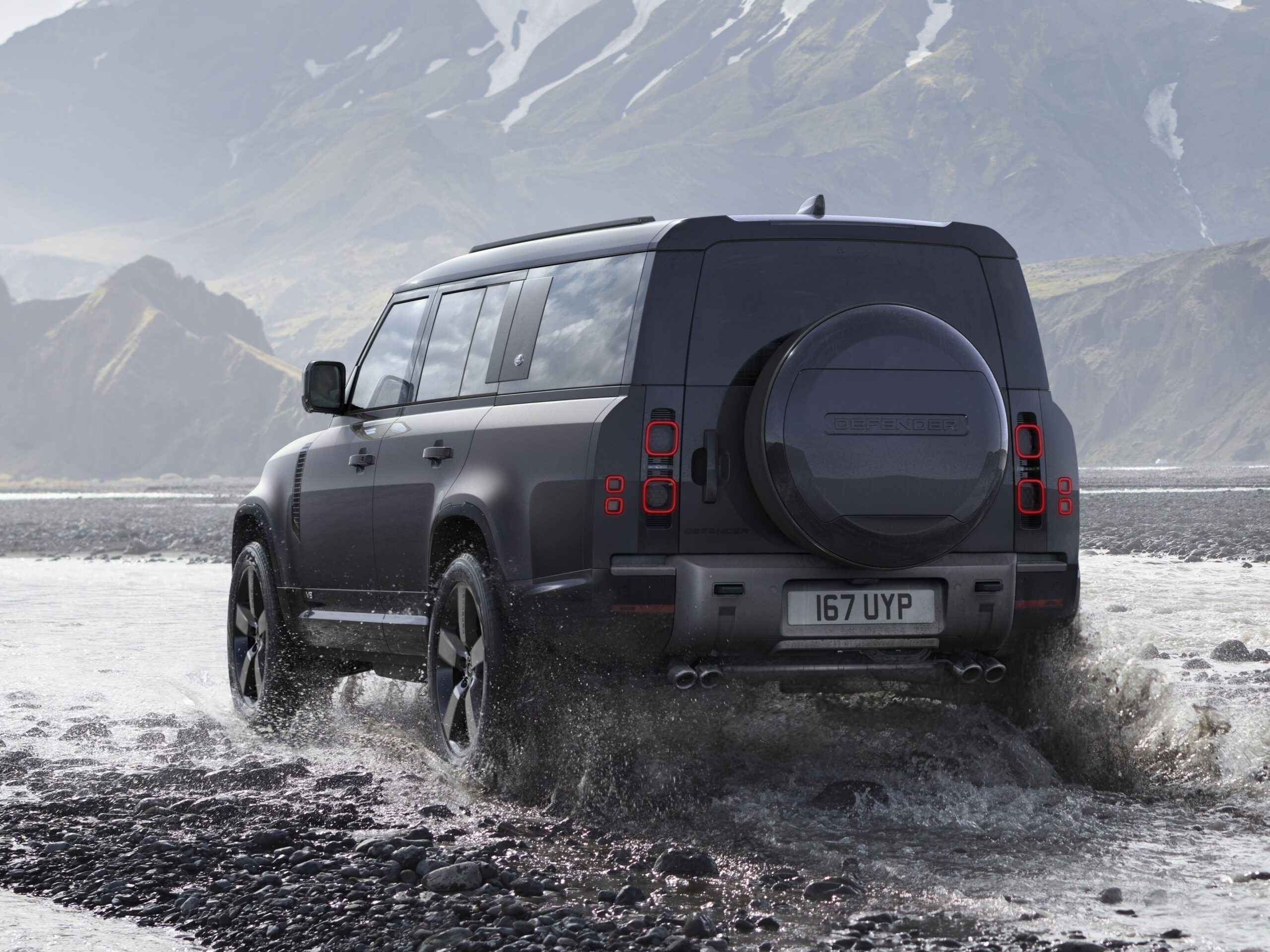 a black land rover defender 110 V8 driving through water next to a mountain