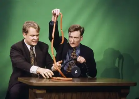 two men behind desk, with conan holding a string of sausages in one hand and a steering wheel in the other