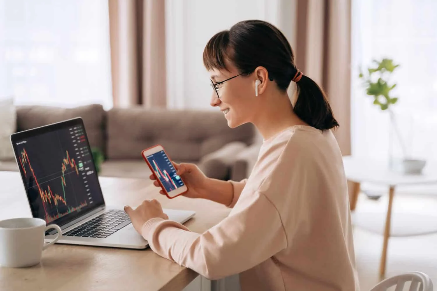 Smiling business woman trader analyst looking at laptop monitor, holding smartphone, wearing earphones. Investor broker analyzing indexes, trading online investment data on stock market graph at home